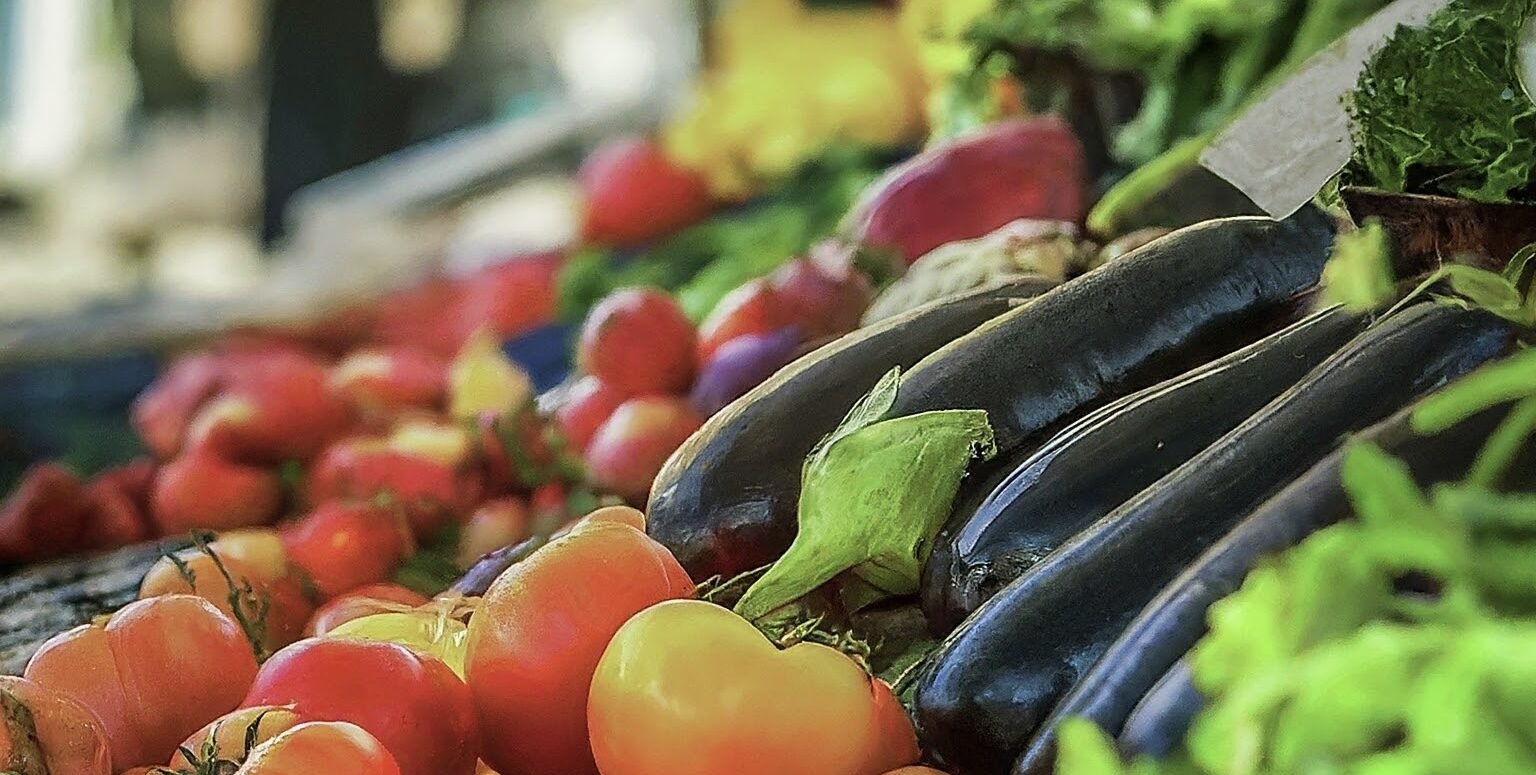 Plan rapproché de poivrons et d’aubergines sombres au milieu d’autres légumes aux couleurs vives, sur fond flou.