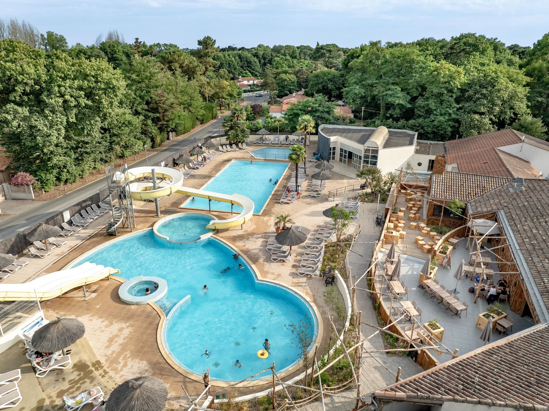Vue aérienne d’une piscine extérieure d’hôtel avec plusieurs bassins, toboggan aquatique, transats et parasols, entourée de bâtiments et de végétation verdoyante.